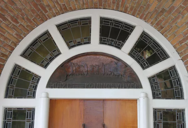Afbeelding uit: juli 2019. Boven de deur bevindt zich een houttableau met daarop de tekst Huize Koningsvijver en een geïdealiseerde voorstelling van het huis.