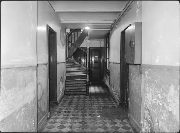 Afbeelding uit: 1959. De hal. Links, rechts en achter voordeuren van woningen; in het midden de trap naar de verdiepingen.
Bron afbeelding: SAA, bestand 5293FO006739.