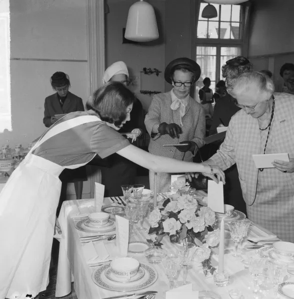 Afbeelding uit: mei 1965. Tafeldekwedstrijd. Met hoed mevrouw Van Hall, echtgenote van de burgemeester; rechts de echtgenote van de ambassadeur van het VK.