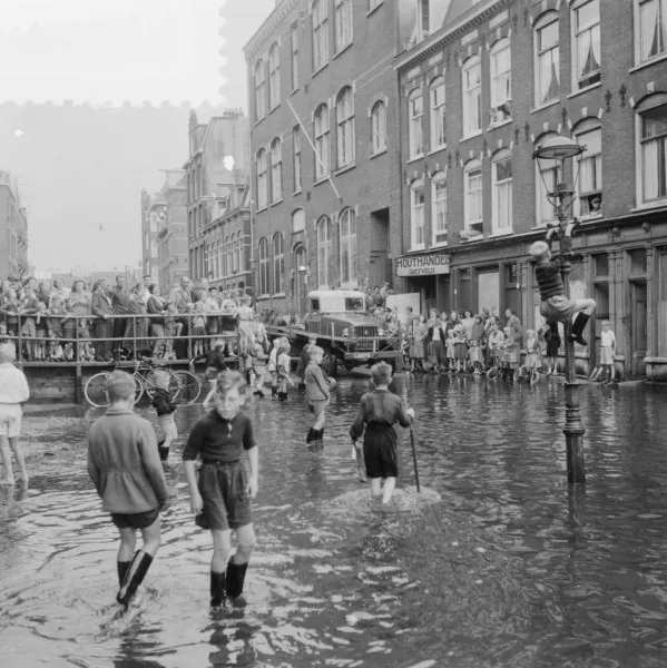 Afbeelding uit: augustus 1951. Kinderen spelen in het water nadat de laaggelegen straat door hevige regenval was ondergelopen. De school is het hoge gebouw.