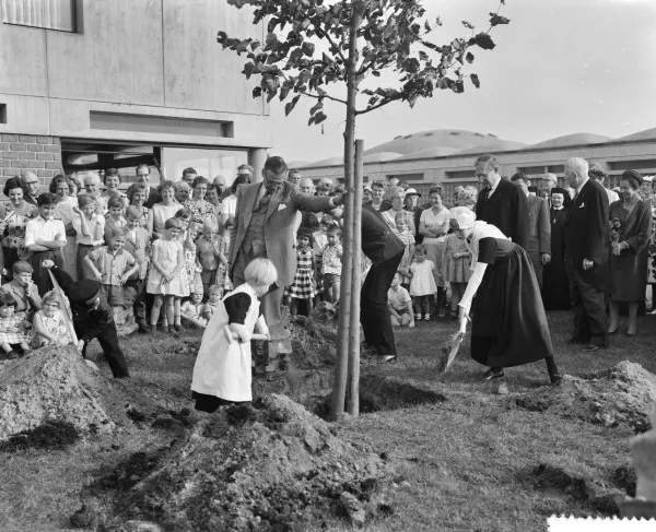 Afbeelding uit: september 1961. Bij de officiële opening werd een boom geplant. Burgemeester Van Hall houdt de boom vast terwijl in traditionele weeshuiskleding gestoken kinderen de kuil dichtgooien.