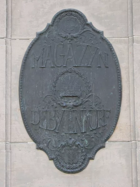 Afbeelding uit: april 2020. Een van de twee plaquettes naast de ingang aan het Damrak. De letters SPG staan voor Simon Philip Goudsmit, de stichter van het winkeltje aan de Nieuwendijk.
