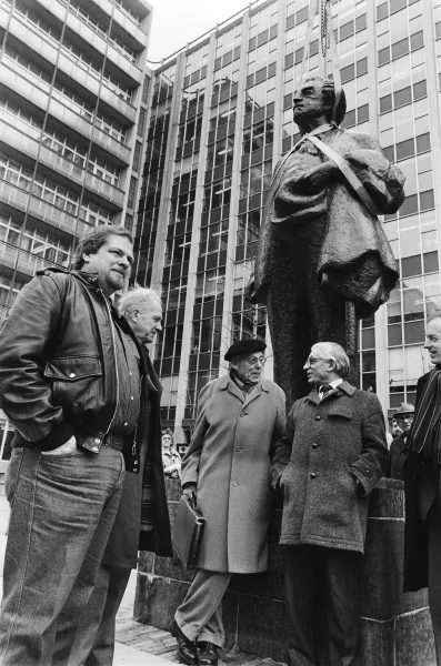 Afbeelding uit: maart 1982. Terug op de sokkel voor het Wibauthuis. Links wethouder Jan Schaefer, met pet beeldhouwer Han Wezelaar, en rechts van hem burgemeester Wim Polak.