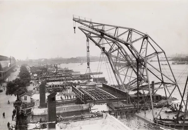 Afbeelding uit: juli 1931. Het beweegbare deel van de brug was gemaakt op de werf van de Amsterdamse Droogdok Maatschappij, de ADM. Hier leggen drijvende bokken van de ADM het op de landhoofden.
Bron afbeelding: SAA, bestand OSIM00004004405.