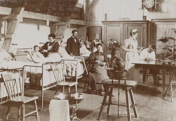 Afbeelding uit: 1890. Een vermoedelijk geënsceneerde foto van een ziekenzaal. Een zuster doet iets met het oor van een jongen, een arts neemt de pols op van een vrouw.
Bron afbeelding: SAA, bestand OSIM00002002257.