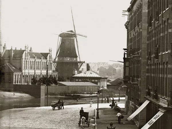 Afbeelding uit: 1890. Uitsnede van een foto uit 1890. Links van het Armenverzorging -gebouwtje staat een school, waar nu de brandweerkazerne is. Erachter molen De Victor, afgebroken in 1898. Op de voorgrond de Nassaukade.