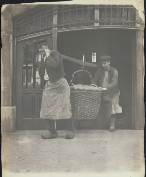 Afbeelding uit: circa 1910. Sjouwers dragen een ogenschijnlijk zware last naar buiten.