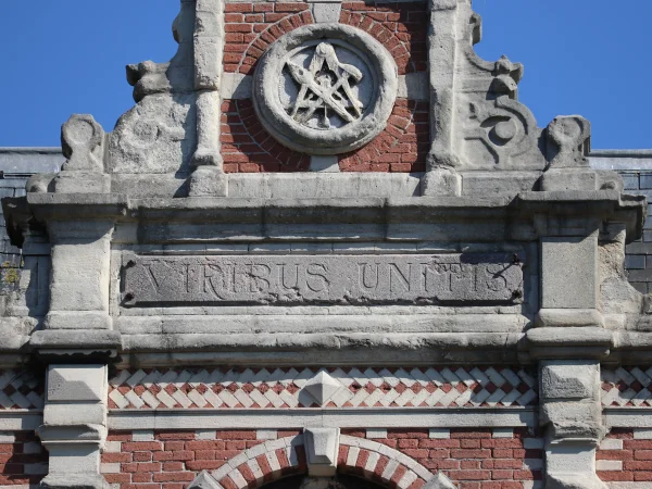 Afbeelding uit: augustus 2024. Bovenin een medaillon met daarin gereedschap zoals passer en hamer. Daaronder de tekst Viribus Unitis: met vereende krachten.