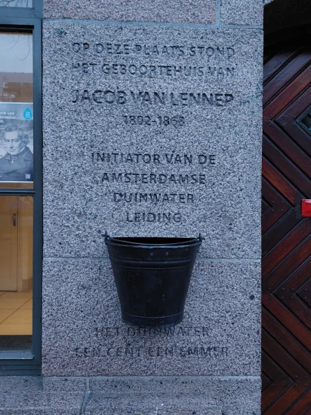 Afbeelding uit: december 2024. Een emmer en een inscriptie: "Op deze plaats stond het geboortehuis van Jacob van Lennep 1802-1868 Initiator van de Amsterdamse Duinwaterleiding [emmer] Het duinwater / een cent een emmer". Geplaatst circa 2024.