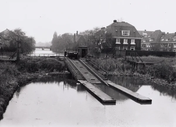 Afbeelding uit: december 1953. Achter de huizen (rechts) lag van 1925 tot 1965 een dam in de Boerenwetering. Die vormde de scheiding tussen water op Amstel- of stadspeil en water op het lagere polderpeil. Boten van tuinders werden met een elektrische overhaal over de dam getrokken.
Bron afbeelding: Stadsarchief.