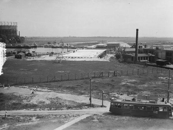 Afbeelding uit: juni 1953. Gezien naar het zuiden. Links een gashouder van de Zuidergasfabriek; rechts een deel van het fabriekscomplex van de ATEK, de Amsterdamse Tricot- en Kunstzijdefabrieken. Op de voorgrond staat een tram van lijn 25 bij de standplaats.
