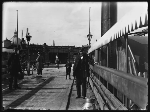 Afbeelding uit: september 1897. De veerboot van de De Ruijterkade naar het Tolhuis aan de Buiksloterweg. Het was een kettingpont, een pont die langs een ketting in het water wordt getrokken. Op de achtergrond is de westelijke onderdoorgang van het Centraal Station te zien.