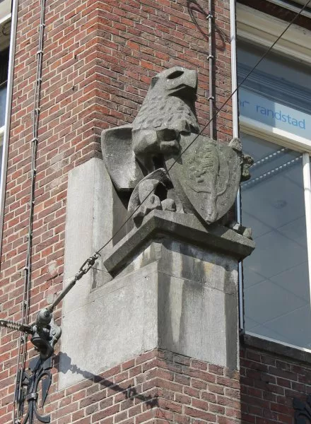 Afbeelding uit: mei 2012. Hoek Damrak. Een griffioen met in zijn klauwen het wapenschild van De Nederlanden. Hij staat op wat met enige goede wil een steunbeer genoemd kan worden, een constructie-element uit de gotiek.